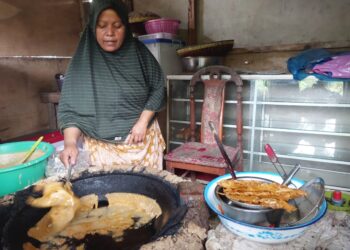 Seorang pedagang gorengan sedang memasak dengan menggunakan wajan besar di dapur sederhana