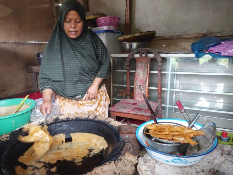 Seorang pedagang gorengan sedang memasak dengan menggunakan wajan besar di dapur sederhana